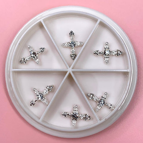 Against a pink backdrop, a round white container showcases the Silver Cross Charm for Nails Art Designer by Dynamic Nail Supply, divided into six sections. Each compartment contains a beautifully arranged rhinestone-studded silver cross charm. These sparkling charms catch the light, highlighting their intricate design and making them perfect for any nails art designer.