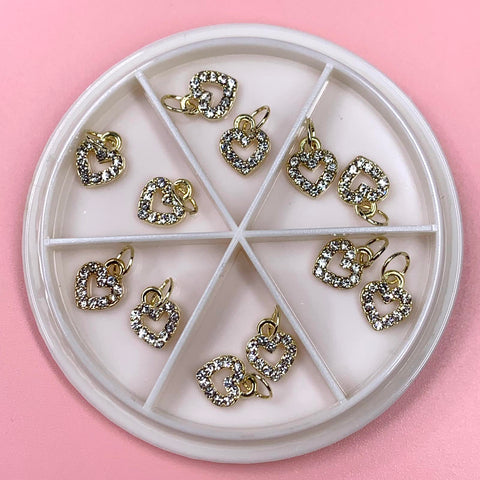 Dynamic Nail Supply's Heart Shaped Dangles Nail Charms set features a round white container that elegantly holds twelve heart-shaped charms. Each gold charm is beautifully bordered with sparkling rhinestones. The tray is divided into six sections, each showcasing two charms against a soft pink background.
