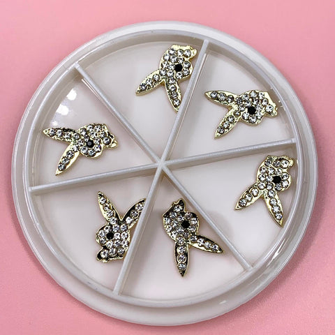 A round white plastic container with six compartments houses Gold Playb*y Bunny Charms for Nail Art Designer from Dynamic Nail Supply, shaped like stylized animals and adorned with small sparkling rhinestones. The charms are set against a soft pink surface, creating an elegant contrast.