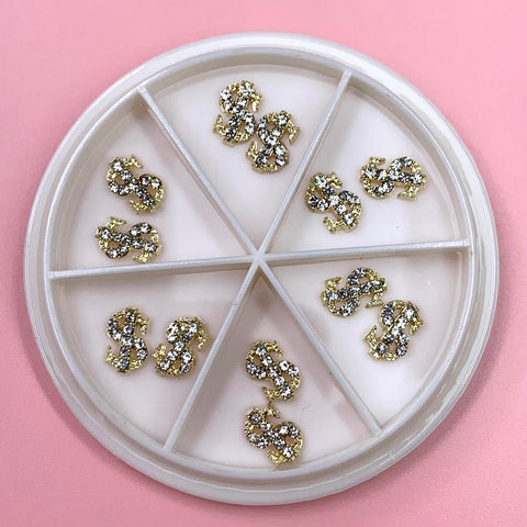 Product Description: A round, white storage case features six compartments, each containing a single gold-colored dollar sign charm adorned with tiny rhinestones. Ideal for nail art enthusiasts, these Gold Dollar Sign Charms for nail art design from Dynamic Nail Supply are beautifully displayed on a pastel pink background.