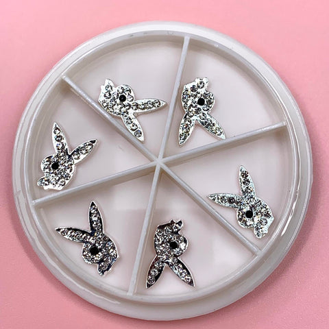 A round white container with six compartments showcases the Silver Playb0y Bunny Charms, set against a light pink background. Ideal for any Nail Art Designer, these charms feature stylized rabbit heads with long ears and small black eye details, adding a chic touch, all sourced from Dynamic Nail Supply.