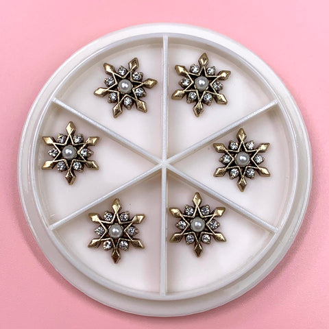 A circular white container featuring six compartments, each containing an Antique Star Flowers Snowflake Charm. These detailed designs, embellished with small pearls and crystals, are perfect for any Nails Art Designer. The pink background from Dynamic Nail Supply enhances the beauty of these decorative pieces.