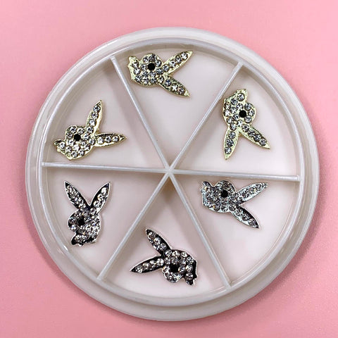 Displayed against a pink background, the 6-grid-wheel box from Dynamic Nail Supply's "3 Silver 3 Gold Playb0y Bunny Charms for Nail Art Designer" showcases sections filled with rhinestone-studded bunny-shaped charms. The bunnies come in various colors, with silver charms and gold stones elegantly arranged throughout the compartments.