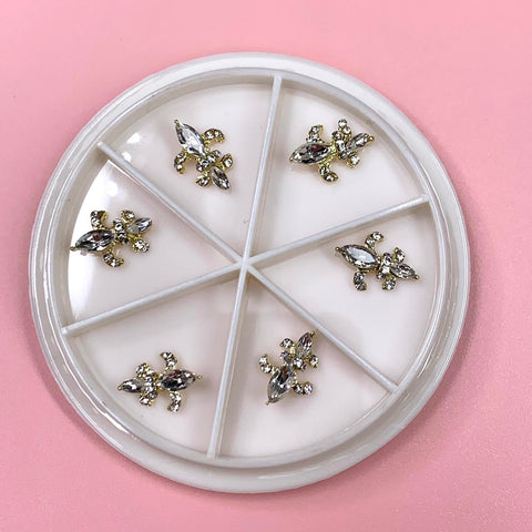 A white round container from Dynamic Nail Supply showcases six compartments against a pink backdrop, each filled with gold Saint Fleur De Lis charms, the Louisiana symbol, embellished with small sparkling rhinestones. The neatly organized and evenly distributed charms bring a touch of elegance to any nail design collection.