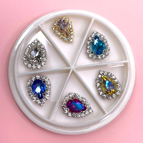 The Heart Shaped Rhinestones Nail Charms Set from Dynamic Nail Supply features a white circular divided container holding six teardrop-shaped gems. Each gem is embellished with heart-shaped rhinestones in silver, gold, blue, yellow, purple, and red hues against a pink background—an eye-catching accessory for any nail art enthusiast.