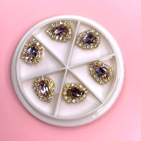 Dynamic Nail Supply presents the Rose Gold Heart Rhinestone Nail Charms, featuring a white circular container with five compartments. Each compartment houses a teardrop-shaped gemstone in a pale purple hue, elegantly accented by rose gold heart rhinestones on a light pink background. The gems are symmetrically arranged within the circle, creating an exquisite and sophisticated design.