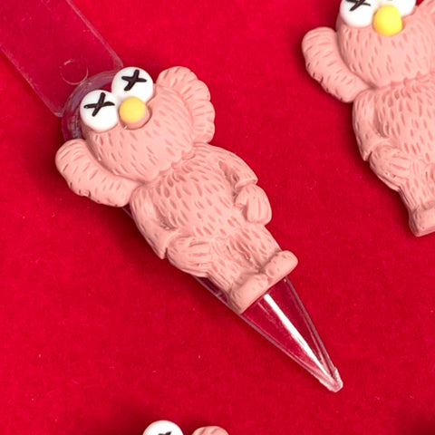 A close-up photo features an adorable clay figure from the Dynamic Nail Supply's 6 pcs KAWS Nail Designer Charms (Pallet Slipper Pink) set. This small creature with X-shaped eyes, ears, and a tail is affixed to a clear, pointed base. The backdrop is a vibrant red, with other figures from the set partially visible around it.