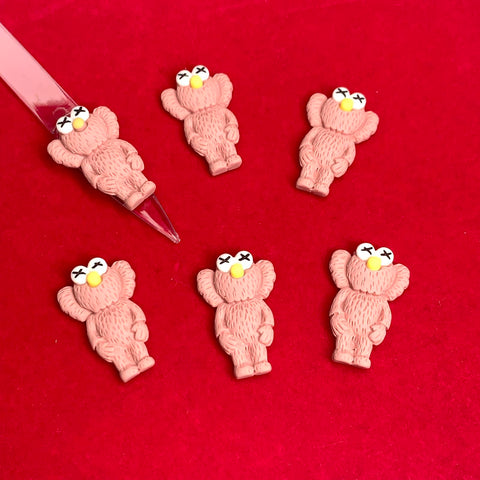 Six KAWS Nail Designer Charms in Pallet Slipper Pink, featuring X-shaped eyes and yellow noses with a fluffy texture, are arranged on a red background. A clear tool from Dynamic Nail Supply points at one of the charms.