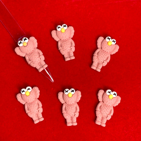 Six small, teddy bear-like figures with exaggerated ears, designed in a pallet slipper pink color and featuring X eyes and yellow noses, are arranged on a vibrant red background. One of the figures is pointed at with a transparent tool. These playful charms resemble the KAWS Nail Designer Charms by Dynamic Nail Supply, complete with furry textures.