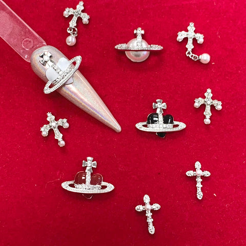 Displayed on a red background is an array of small, decorative jewelry pieces from Dynamic Nail Supply. Among the collection are silver cross crucifix charms embellished with clear stones and small pearl-like accents, as well as space-themed planet shapes with stone-encrusted rings. Also featured is a shimmering nail art sample that showcases a planetary design.