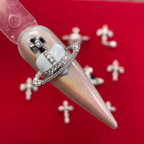 A pointed nail design features a stunning application of Nail Art with a 3D silver charm depicting a planet, adorned with both a ring and cross. Set against a red backdrop, the celestial theme is enhanced by the addition of similar Silver Cross Crucifix Space Planet Charms from Dynamic Nail Supply, creating an exquisite and iridescent look.