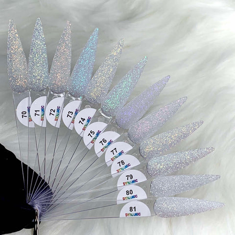 A stunning fan display of nail art swatches on long, pointed tips showcases various shades featuring Dynamic Nail Supply's Iridescent Loose Glitter in non-toxic, glittery white and silver. Each swatch is labeled with a number from 70 to 81 and is set against a background of soft, white fuzzy fabric.