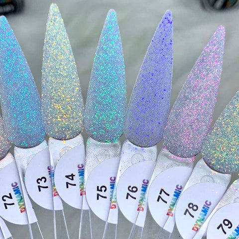 Close-up of a display featuring nail polish samples numbered 73 to 79. Each long, pointed tip showcases a unique glittery shade ranging from silver to light purple, exhibiting diverse holographic effects and loose glitter. The tips are mounted on clear sticks labeled "Dynamic Nail Supply." Ideal for creating stunning, non-toxic nail art using Iridescent Loose Glitter (Super-Fine Size). The background is blurred.