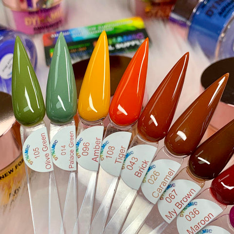 A close-up shot features eight pointed sample sticks from the "2024 Fall Acrylic Powder Collection (part 2) Newest" by Dynamic Nail Supply, arranged in a fan. The shades vary from olive green to maroon, each labeled with stickers: Olive Green, Pale Green, Amber, Tiger, Brick, Caramel, My Brown, and Maroon. In the background are various brightly colored polish bottles that appear blurred.
