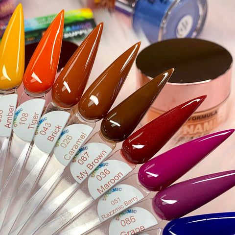 A fan-shaped display of elongated, pointed nail color samples showcases Dynamic Nail Supply's 2024 Fall Acrylic Powder Collection (part 2) Newest. The collection features a range of shades from amber, orange, brown, and maroon to deep purple. Each sample is labeled with its color name and number, with a jar of acrylic powder visible in the background.