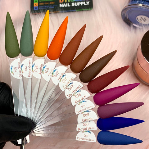 A fan display of long, pointed nail tips showcases various matte shades from the 2024 Fall Acrylic Powder Collection (part 2) Newest by Dynamic Nail Supply, each labeled with names like Olive Green, Amber, and Egyptian Blue. Held on a furry white surface by a gloved hand, these nails promise to be as stunning as they are long-lasting.