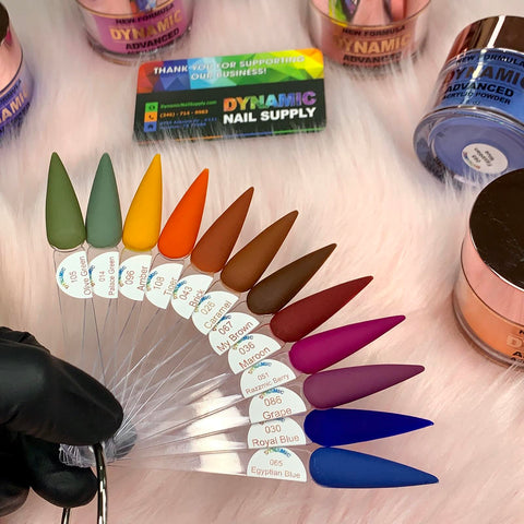 A display of nail polish samples on fan-shaped sticks features ten vibrant colors: olive green, mint green, mustard yellow, bright orange, reddish brown, medium brown, light brown, beige, mauve, and deep blue. In the background is a Dynamic Nail Supply sign promoting its 2024 Fall Acrylic Powder Collection (part 2) Newest for long-lasting nails.