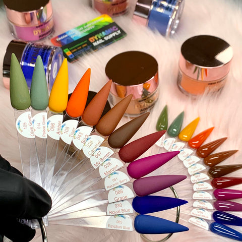 A display of vibrant nail polish samples from Dynamic Nail Supply's 2024 Fall Acrylic Powder Collection (part 2) Newest is artfully arranged on a fan ring, highlighting a variety of shades including greens, oranges, and blues. In the background, nail supplies such as jars and containers are visible alongside a gloved hand holding the long-lasting acrylic powders against a plush white surface.