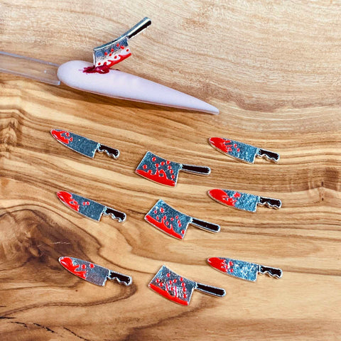 A wooden surface showcases a set of 10 charms from Dynamic Nail Supply, designed for Halloween nail art. These 3D charms are shaped like small cleavers with shiny silver blades and black handles. Red paint mimicking blood splatters adds to their spooky theme, especially seen on one charm which is attached to a nail holder with the red paint dripping down.