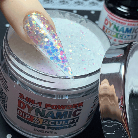 A close-up of a finger with a glittery, multicolored, stiletto-shaped nail is dipping into a jar labeled "White Holographic Glitter Nail Powder - 327 Galaxy" by Dynamic Nail Supply. The powder is white with iridescent sparkles. In the background, there is a bottle showcasing stunning nail designs and bearing the Dynamic label.