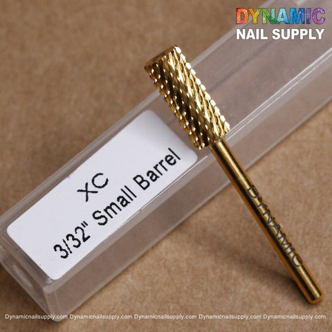 A close-up of the gold-colored cylindrical drill bit, labeled with "DYNAMIC," is placed next to a white box marked "XC Small Barrel 3/32." The scene, set against a light brown surface, includes Dynamic Nail Supply's website details along the bottom edge.