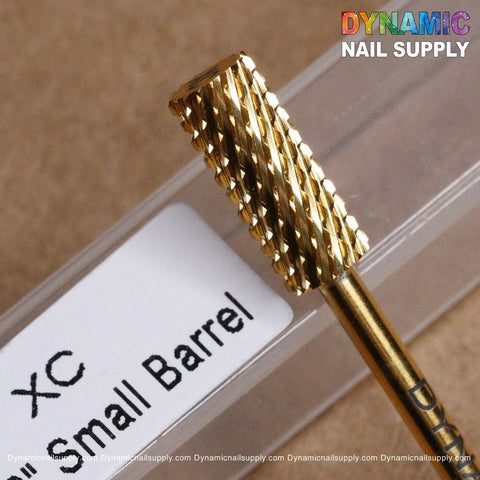 Close-up of the Dynamic Nail Supply XC Small Barrel - Dynamic Drill Bit in a gold-colored carbide finish. The textured surface makes it ideal for filing, and it's showcased on a clear plastic container featuring Dynamic Nail Supply's branding and website URL in colorful lettering in the background.